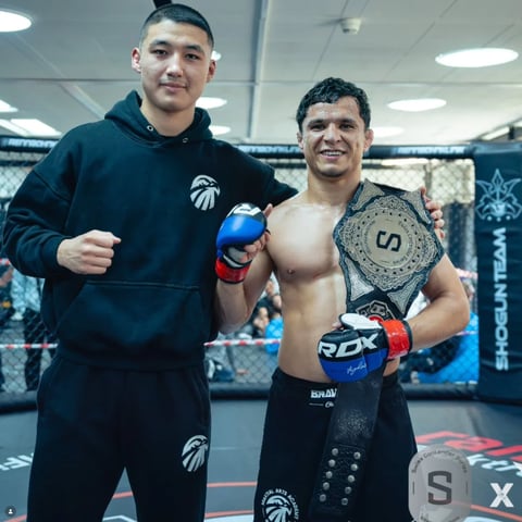 Two male fighters posing in an MMA octagon cage, one wearing a navy hoodie and the other shirtless with a championship belt
