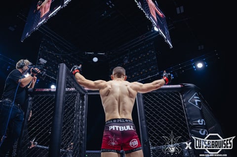 Shirtless MMA fighter stands in octagon cage with arms raised, facing audience during professional fighting event
