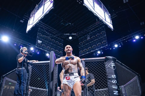 Muscular MMA fighter in cage with arms spread, ring announcer holding microphone inside octagon arena