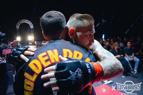 Two men embracing in a boxing ring during a match, with crowd and bright lights visible in the background.