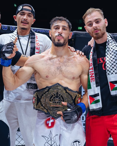 Shirtless MMA fighter wearing championship belt poses with two men in corner team gear at boxing event