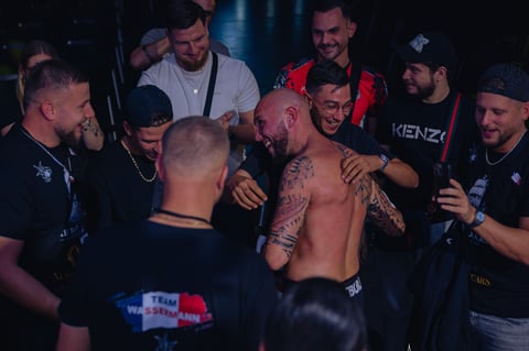 A shirtless man with tattoos celebrates with a group of friends in a dark venue or club setting