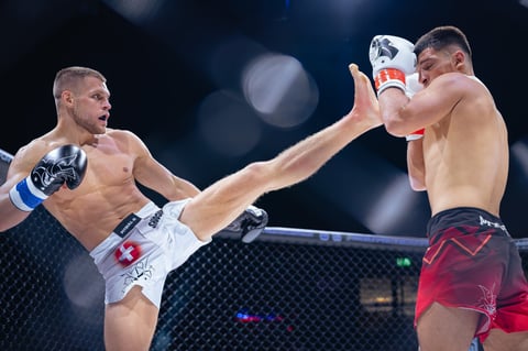 Two shirtless MMA fighters in an octagon cage, one throwing a high kick at his opponent during a professional mixed martial arts match.
