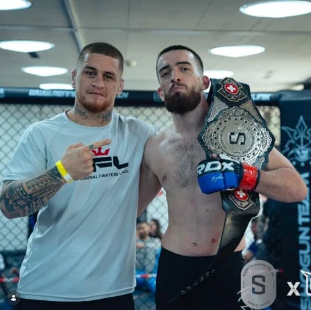 Two men posing in an MMA gym, one wearing a UFC shirt and the other shirtless holding a championship belt