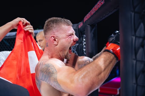 Muscular man with tattoos holding Swiss flag while wearing red boxing gloves in ring
