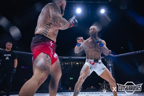 Two shirtless MMA fighters in an octagon cage during a professional mixed martial arts bout with bright stage lights.