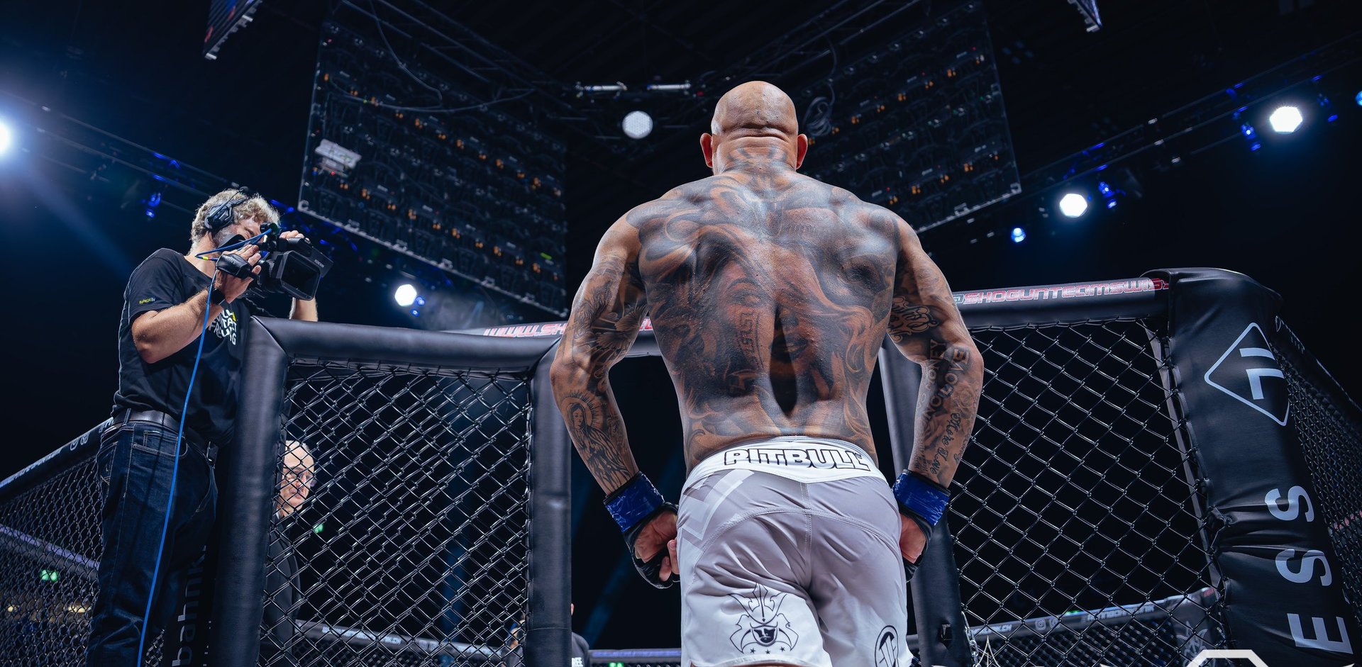 Shirtless fighter standing in octagon cage with referee and crowd lights in background at MMA event