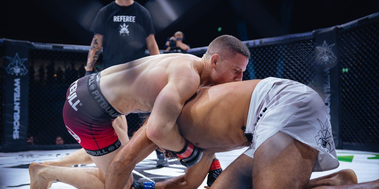 Two MMA fighters grappling during a match inside an octagon cage with referee visible in background