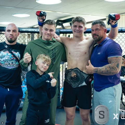 Young boxer and four men posing together in an MMA gym by the octagon cage, celebrating after training or a match